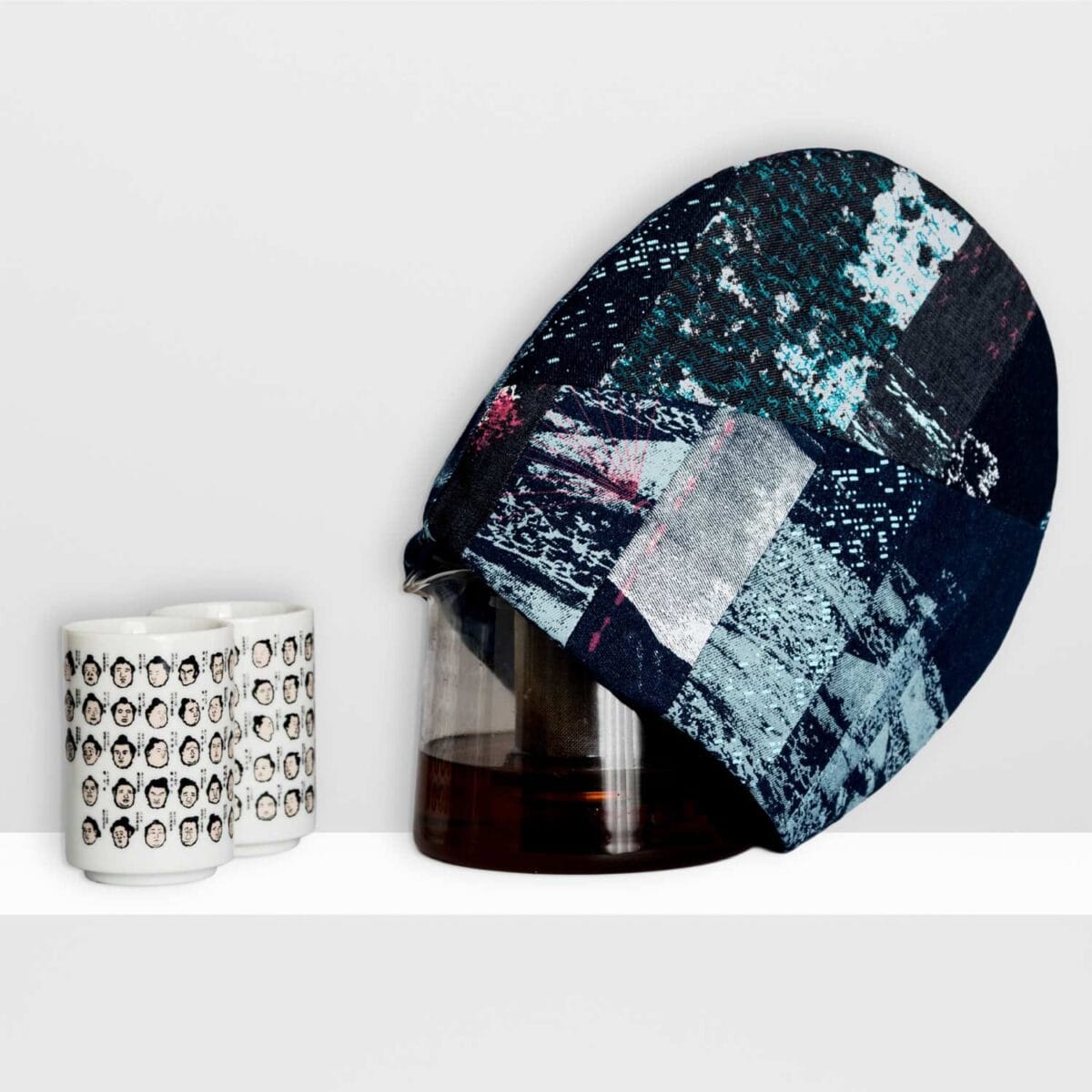 Composition of two ceramic mugs with faces of sumo wrestlers and a tea pot partly covered by a tea cosy by MIN-studio made of screen-printed denim with layered moon surface textures in blue, grey, and neon pink inks, featuring edgy, grunge-inspired abstract patterns.
